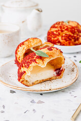 Small cakes profiterole choux pastry with custard, strawberry jam and red icing on a white concrete background.