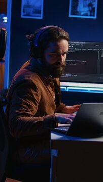 Developer focused on typing complex code in neon lit home office, listening music and developing software application. IT expert wearing headphones, solving coding project tasks on laptop, camera B