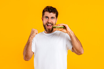 joyful guy biting cheeseburger and gesturing yes on yellow background