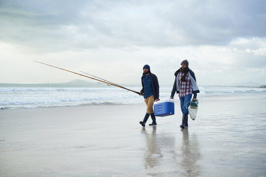 Winter, fishing and men walking on beach together with cooler, tackle box and holiday conversation. Ocean, fisherman or friends with rods, bait and tools in nature on morning vacation with cloudy sky