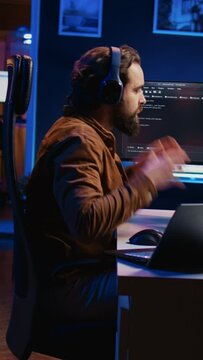 Technician focused on typing complex code in neon lit home office, developing software application. It specialist removing headphones to better concentrate on difficult task, camera B