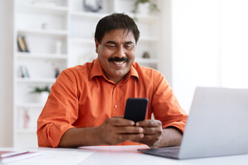 Fototapeta premium Indian middle aged businessman texting on cellphone and using laptop