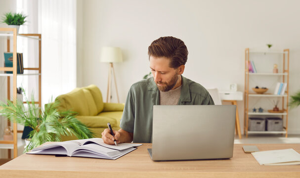 Portrait of a young business man in casual clothes working on a laptop computer at office or at home and writing at the desk making notes or calculations. Remote and freelance work concept.