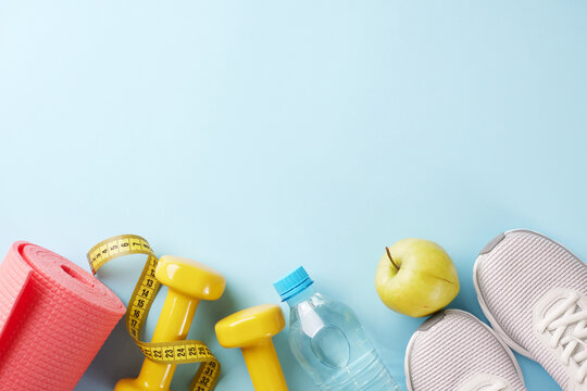 Push your limits: transform your body through active living. Top view shot of trendy sneakers, yellow dumbbells, measure tape, yoga mat, water, apple on pastel blue background with promo area