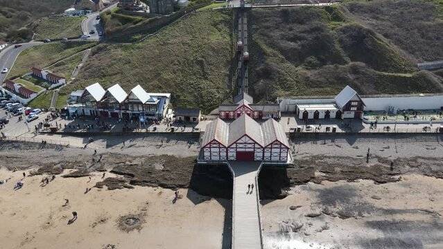 UK: Saltburn-by-the-Sea: An aerial footage of a sea pier with building area in the background, sandy beach and calm sea water