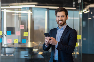 Successful mature businessman smiling with smartphone in hand, standing in a contemporary office setting.