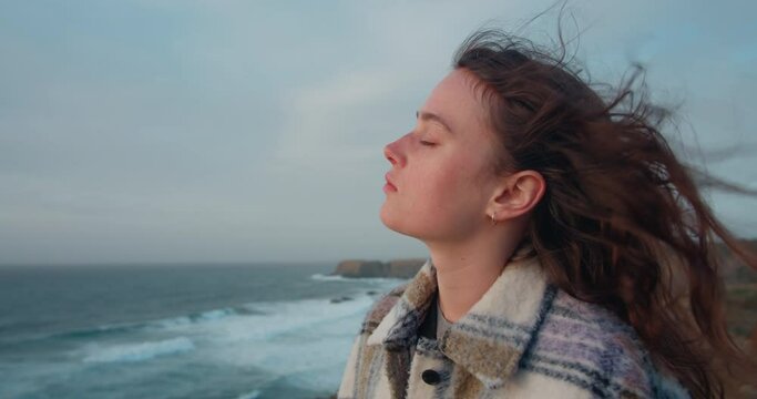 Cinematic portrait of young woman with closed eyes enjoying wind breeze in her hair, deep in thoughts, breathing in fresh ocean air. Concept of freedom and travel mood