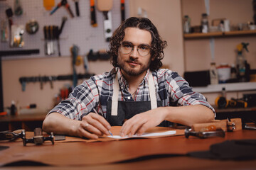 Cobbler Man tailor cuts out blanks for sewing bags or shoes according to pattern made of leather in workshop