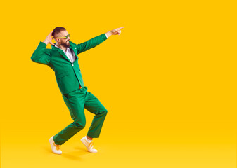 Fototapeta na wymiar Happy man pointing his finger at copy space. Full size side view shot of excited man in emerald green suit and sunglasses having fun and laughing at something on isolated on yellow background