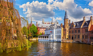 Obraz premium Bruges, Belgium. Ancient medieval european city. View at tower and vintage building on the banks of Rozenhoedkaai channel river. Panoramic view with blue sky clouds. Famous tourist attraction.