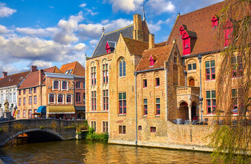 Fototapeta premium Bruges, Belgium. Ancient medieval european city. View at tower and vintage building on the banks of Rozenhoedkaai channel river. Old stone Bridge. Panoramic view with blue sky clouds. Famous tourist a