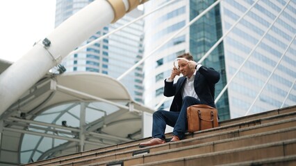 Smart business man wear headphone while sitting at stair in urban city and listen to relaxing music. Happy male leader using headset and moving to song in lively mood surrounded by skyscraper. Urbane.