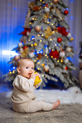 Small child sits on the floor near the Christmas tree and drinks juice from bottle.