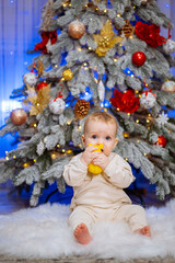 Small child sits on fur rug in front of decorated Christmas tree and eats tangerine.