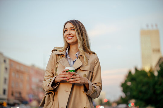 A happy woman is holding coffee to go on a city street.