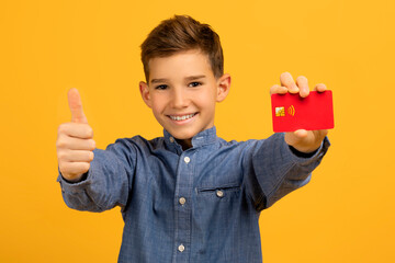 Cheerful teenage boy presenting red credit card and showing thumb up gesture