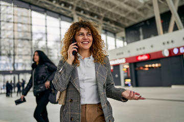 Amidst the bus station's activity, a joyous 30s woman with curly hair strolls, engaged in a phone...