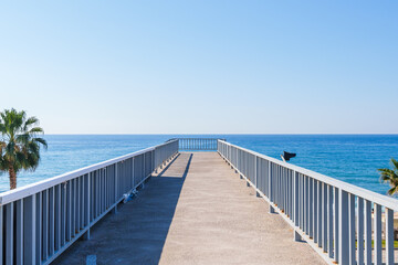 Fototapeta premium Empty Stone Pedestrian Crosswalk Bridge Over a Highway with a Blue sea and Palm Trees Behind it Under the Clear Sky in a Hot Summer Day