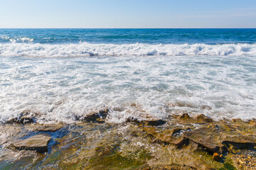 Fototapeta na wymiar Blue Clear Sea Waves Splashing on a Rocky Beach during a Windy Day With Clear Blue sky