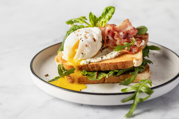 Toasts sandwich with poached egg, arugula, bacon and cream cheese on white plate marble background