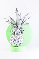 Artistic composition with silver metallic pineappple on light green round podium on white background close up.