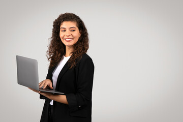 Confident businesswoman in black suit holding laptop with cheerful expression and looking back at free space,