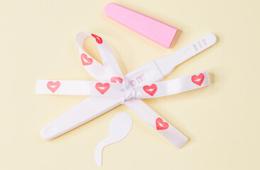 Pregnancy test with heart ribbon and sperm paper on light yellow.