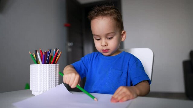 Cute smiling baby in blue t-shirt drawing with green pencil. Kid finishes his picture and puts a pencil into a cup. Blurred backdrop.