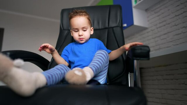 Cute baby boy wants to get up on the rotating chair. Little child with gadget in hands. Low angle view.