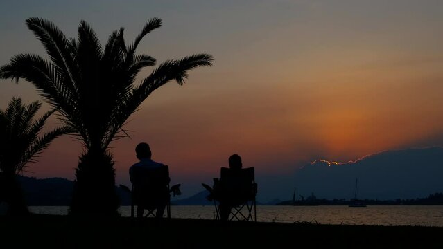 Couple silhouette on empty tropical bay. A view of man and woman silhouette sitting on beach chairs by the waves during tropical summer evening.