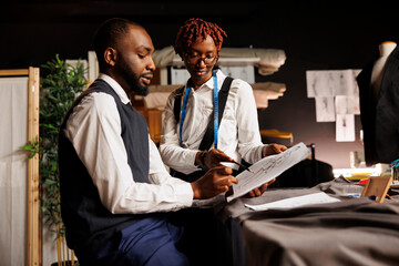 Brainstorming apprentice tailors looking over sketch drawings of bespoke collection imagined by...
