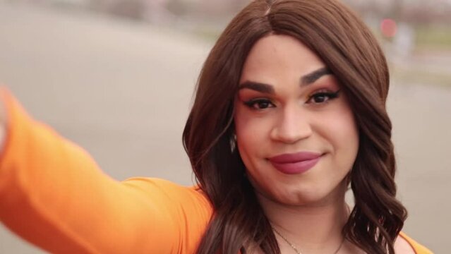 Self-video of trans woman in orange clothes. Latin ethnicity. City outdoors.