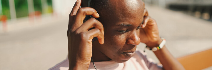Portrait of young african guy relaxes sitting on bench in the port, listens to music in headphones,...