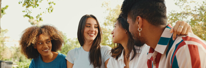 Happy multiethnic young people talking while sitting on park bench on summer day outdoors,...