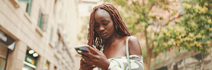 Gorgeous woman with African braids wearing top stands outside on the street and uses mobile phone,...
