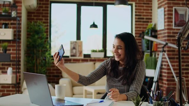 Laughing happy indian woman taking selfie with mobile phone while smiling at home office desk. Person in nice stylish modern house capturing photo using smartphone camera, camera A