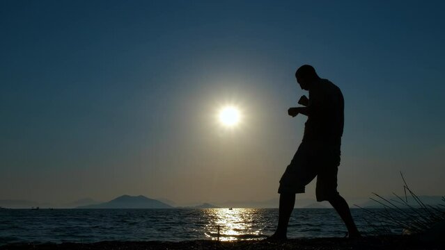 Man silhouette doing shadow boxing. A view of sporty man silhouette practising sport on the empty beach during sunset in summer.