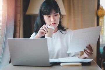 Business analytics concept, A focused Asian businesswoman attentively evaluates a business report while sipping coffee in a cafe with her laptop open.