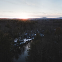 An aerial image taken from an elevated position above the ground. The Nashua River is in the foreground and Mount Wachusett is in the background. Taken as the sun was setting over the forested scene.