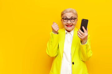 old businesswoman in glasses and formal wear uses smart phone and celebrates victory on yellow...