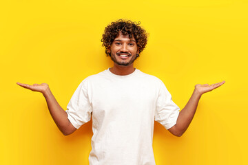 cheerful curly indian man in white t-shirt holds empty hands to sides on yellow isolated...