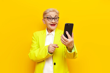 old businesswoman in glasses and formal wear using smartphone and laughing on yellow isolated background, elderly pensioner grandmother in blazer typing and selecting online in mobile