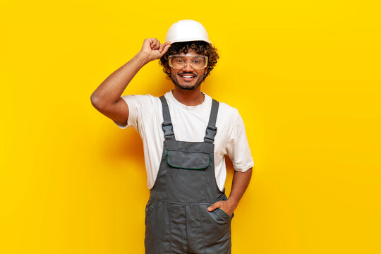 young indian male builder in hard hat and overalls smiling and looking at camera on yellow isolated background, indian foreman in uniform and safety glasses