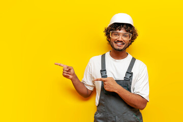 young indian male builder in hard hat and overalls pointing to the side on yellow isolated...