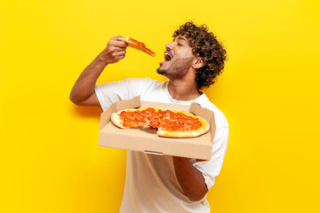 young indian man holding pizza box and eating a slice on yellow isolated background, curly guy student eating and advertising fast food