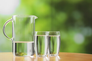 Jug and glasses with clear water on table against blurred green background, closeup. Space for text