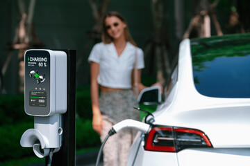 Young woman travel with EV electric car charging in green sustainable city outdoor garden in...