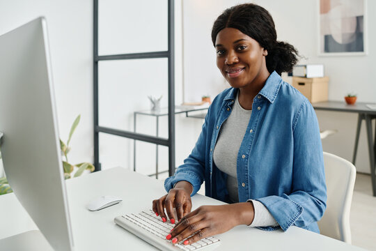 Medium portrait of confident young African American female manager wearing casual outfit smiling at camera while working on computer in modern office