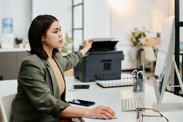 Side view shot of young female secretary printing documents while working on desktop computer in modern office