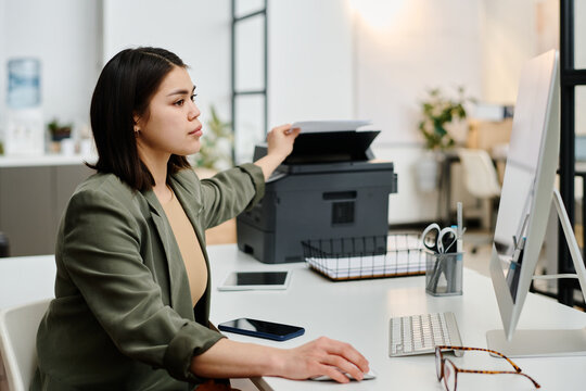 Side view shot of young female secretary printing documents while working on desktop computer in modern office
